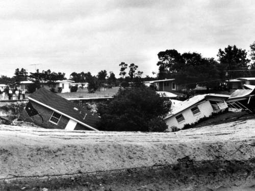 A black and white photo of houses tilted and damaged, surrounded by a large sinkhole and trees in the background.