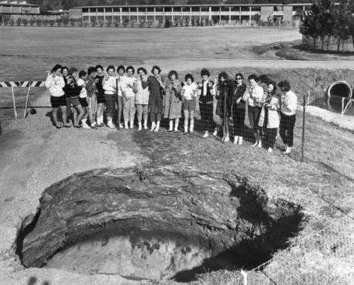 A group of people stands by a large hole in the ground, with a building and grassy field in the background.