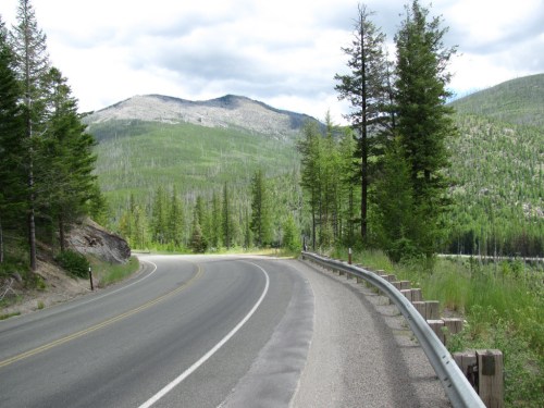 A winding road surrounded by lush green trees and mountains under a cloudy sky.