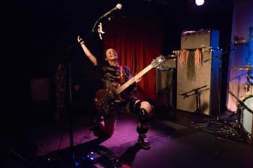 A musician kneels on stage, playing guitar passionately with a microphone in hand, surrounded by instruments and colorful lights.