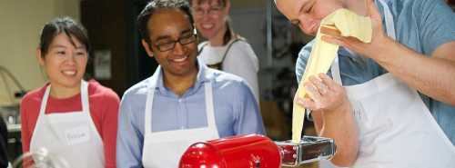 A group of four people in an apron, enjoying a cooking class, with one person stretching dough from a mixer.