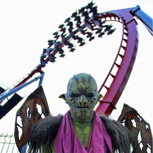 A menacing creature with wings stands in front of a roller coaster, which features a steep loop and riders above.