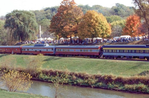 A colorful train travels along a river, with trees in autumn colors and a crowd gathered on a hillside.