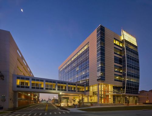Modern glass building at dusk, featuring a sky bridge and illuminated windows, with a clear sky above.