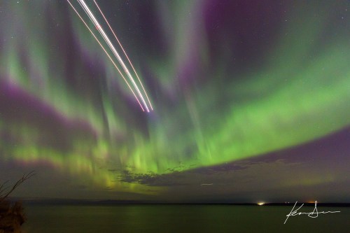 A vibrant display of the northern lights with streaks of light from planes above a calm water surface.