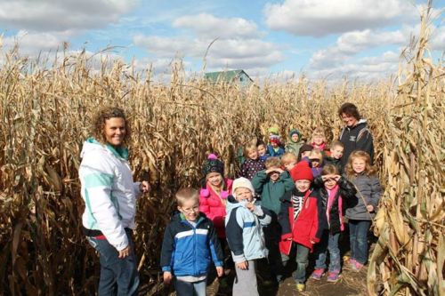 A group of children and adults smiling in a corn maze on a sunny day, surrounded by tall corn stalks.