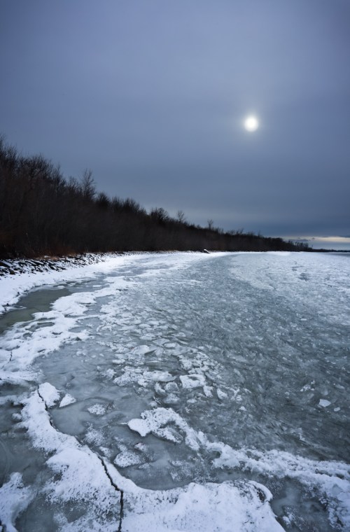 A frozen shoreline under a cloudy sky, with patches of ice and a faint sun in the background.