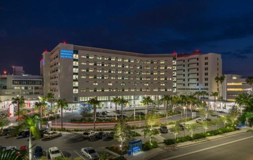 A modern hospital building illuminated at night, surrounded by palm trees and parked cars.