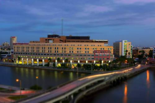 A large hospital building by a river, with city lights and a cloudy sky in the background.