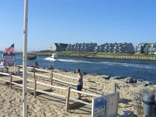 A sandy beach with a boat passing by, surrounded by buildings and a flag waving in the breeze.