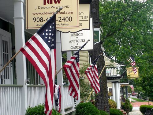 A street scene featuring American flags hanging from buildings, with shops and greenery in the background.