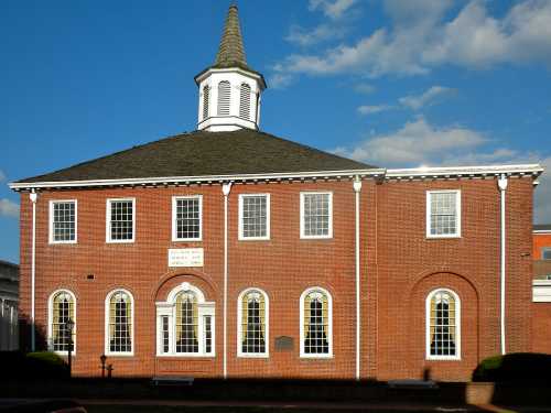 Historic brick building with a steeple, featuring large windows and a plaque, set against a blue sky.