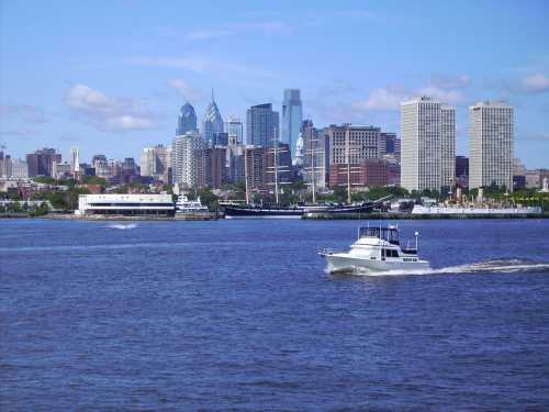 A boat glides across the water with a city skyline in the background under a clear blue sky.