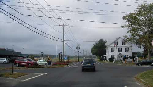 A rural intersection with cars, power lines, and a white building in the background under a cloudy sky.