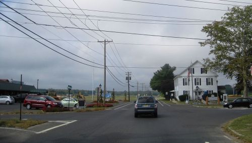 A rural intersection with cars, power lines, and a white building in the background under a cloudy sky.
