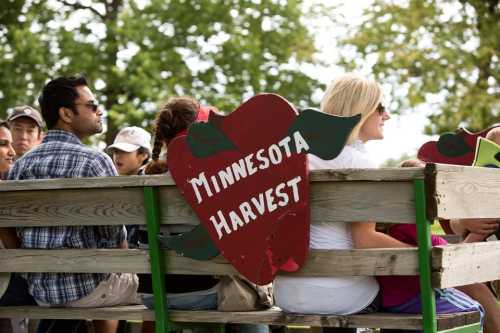 A group of people sitting on a wagon with a sign that reads "Minnesota Harvest" in a sunny outdoor setting.