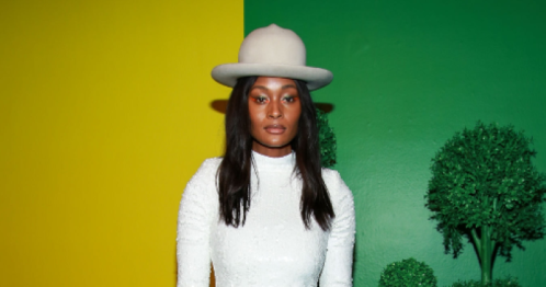 A woman in a white dress and gray hat stands against a vibrant yellow and green background.