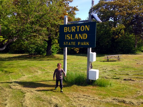 A child stands in front of the Burton Island State Park sign, surrounded by green grass and trees.