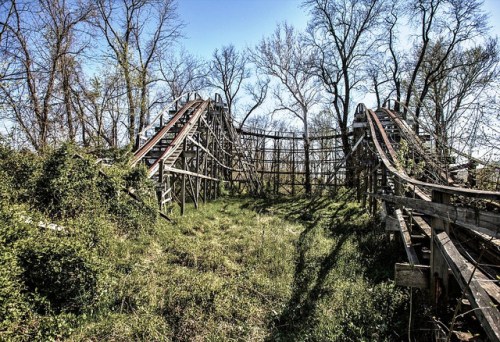 Abandoned wooden roller coaster overgrown with grass and trees under a clear blue sky.