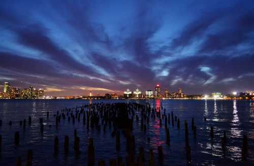 A serene waterfront scene at dusk, featuring silhouetted pilings and a city skyline under a colorful sky.