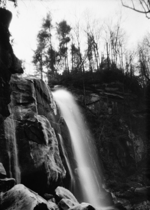 A black and white image of a waterfall cascading over rocks, surrounded by trees and a rugged landscape.
