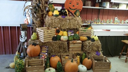 A festive display of pumpkins and gourds arranged on hay bales and wooden crates, with Halloween decorations in the background.