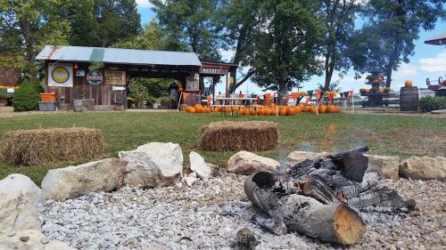 A rustic market with pumpkins, hay bales, and a fire pit, set against a backdrop of trees and blue sky.