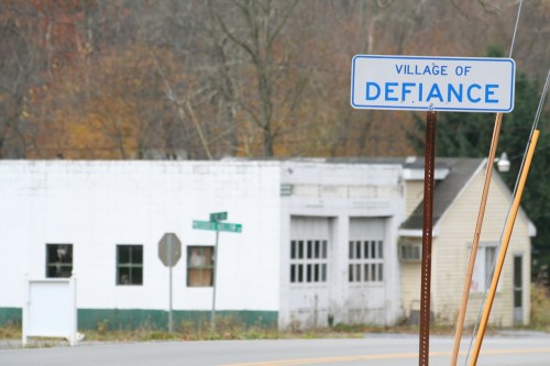 Sign for the Village of Defiance, with a faded building and street signs in the background.