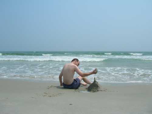 A person sitting on the beach, building a sandcastle near the water's edge on a sunny day.