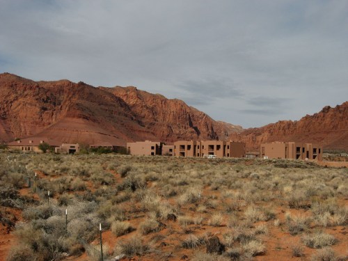 Desert landscape with brown buildings against red mountains and sparse vegetation under a cloudy sky.