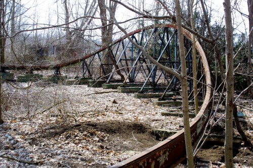 An abandoned roller coaster track overgrown with trees and foliage in a wooded area.