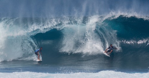 Two surfers riding large waves under a clear blue sky, showcasing dynamic movement and ocean spray.