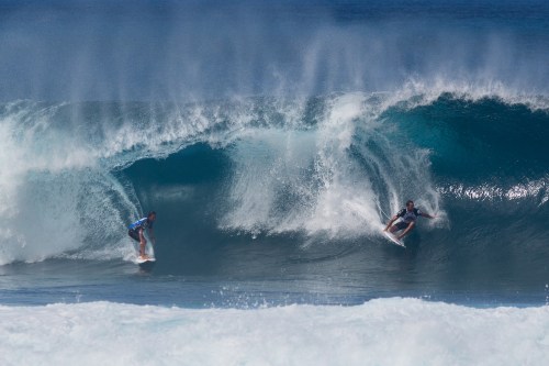 Two surfers riding large waves under a clear blue sky, with splashes of water around them.