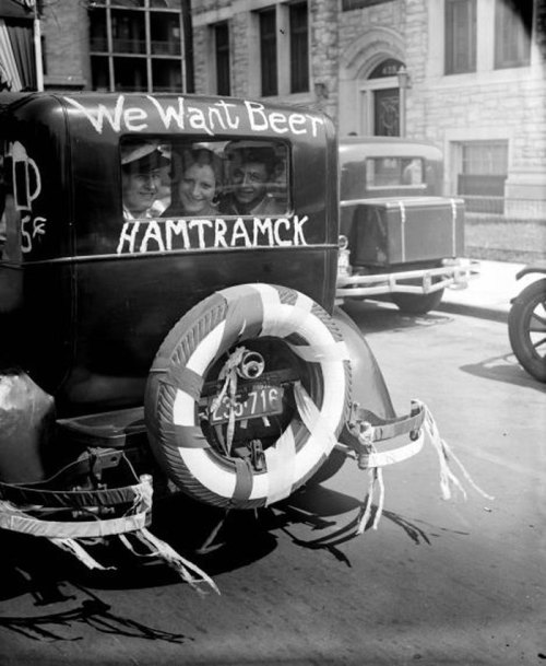 A vintage car with "We Want Beer" and "HAMTRAMCK" painted on the back, featuring smiling passengers inside.