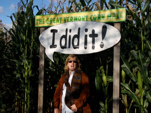 A woman stands in front of a sign that reads "I did it!" at the Great Vermont Corn Maze, surrounded by tall corn stalks.