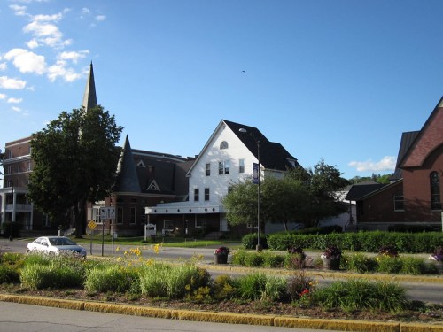 A scenic view of a street with historic buildings, a church spire, and colorful flower beds under a clear blue sky.