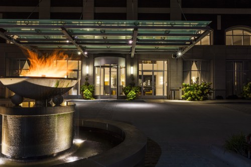 A modern building entrance at night, featuring a lit fountain and lush greenery under a glass canopy.