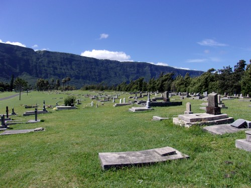 A serene cemetery with gravestones on green grass, set against a backdrop of mountains and a clear blue sky.