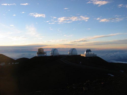 Observatories on a mountain peak at sunset, with clouds below and a colorful sky above.