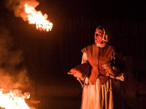 A woman in historical attire holds chickens, standing near a fire at night, with a torch illuminating the scene.