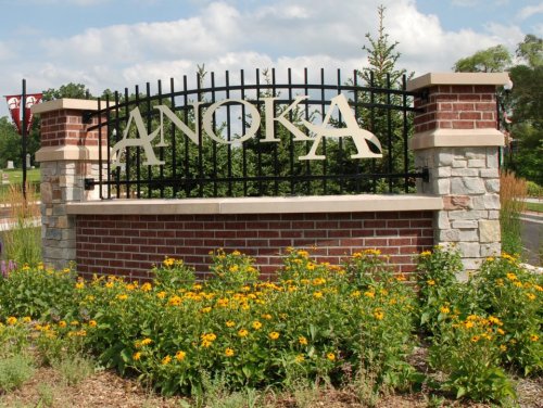 Sign reading "ANOKA" on a decorative stone and metal structure, surrounded by yellow flowers and greenery.