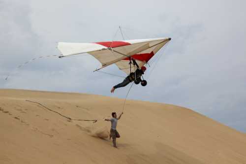 A person launches a hang glider from a sandy dune, while another assists by holding the glider's frame.