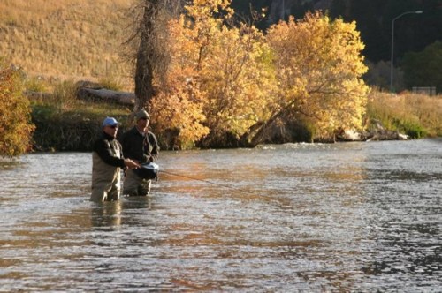 Two anglers stand in a river, casting lines, surrounded by autumn foliage and calm waters.
