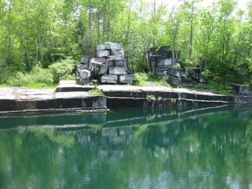 A serene landscape featuring a rocky shoreline with green trees and calm water reflecting the rocks.