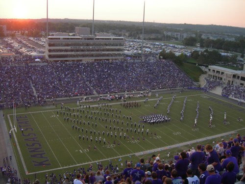 Aerial view of a football stadium filled with fans, featuring a marching band performing on the field at sunset.