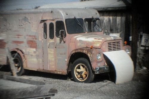 An old, rusty delivery truck with peeling paint, parked beside a wooden structure.