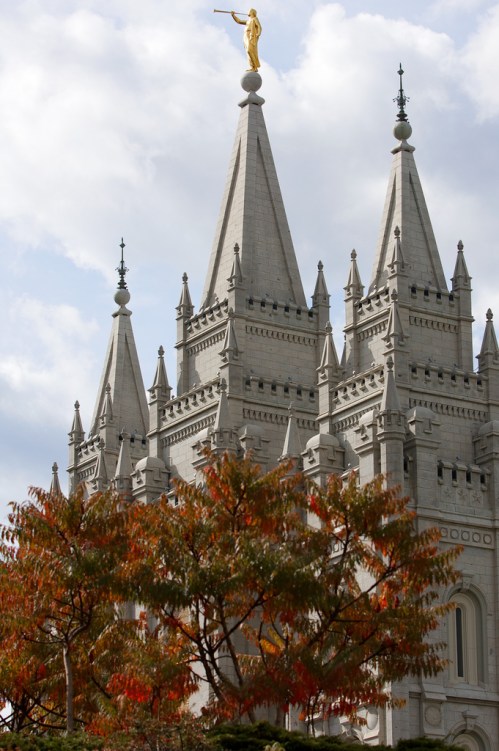 The image shows a tall, ornate building with spires, topped by a golden statue, surrounded by autumn foliage.