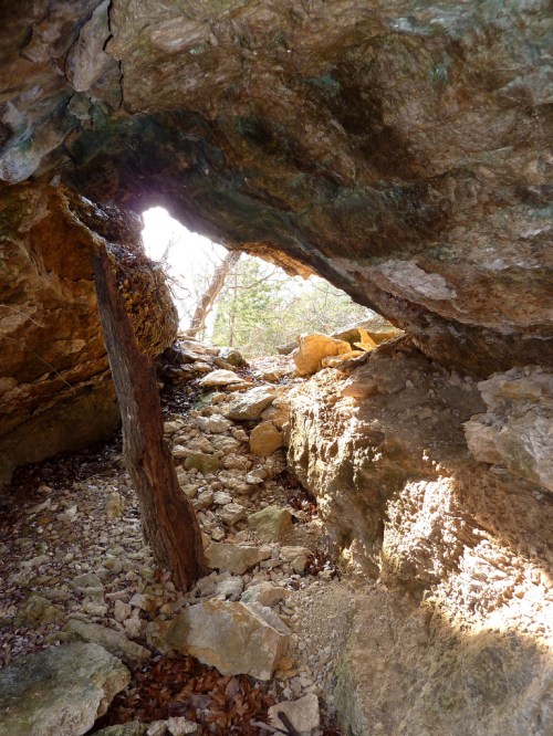 A rocky cave entrance with sunlight streaming through, illuminating the surrounding stones and vegetation.
