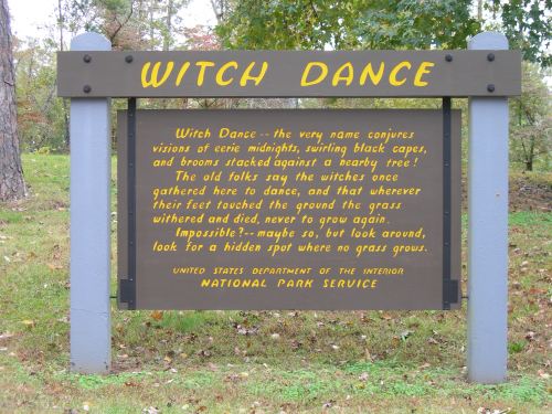 Sign for "Witch Dance" with text about local folklore and a warning about the grass. Surrounded by trees.