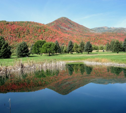 A serene landscape featuring a calm pond reflecting colorful autumn foliage and mountains under a clear blue sky.